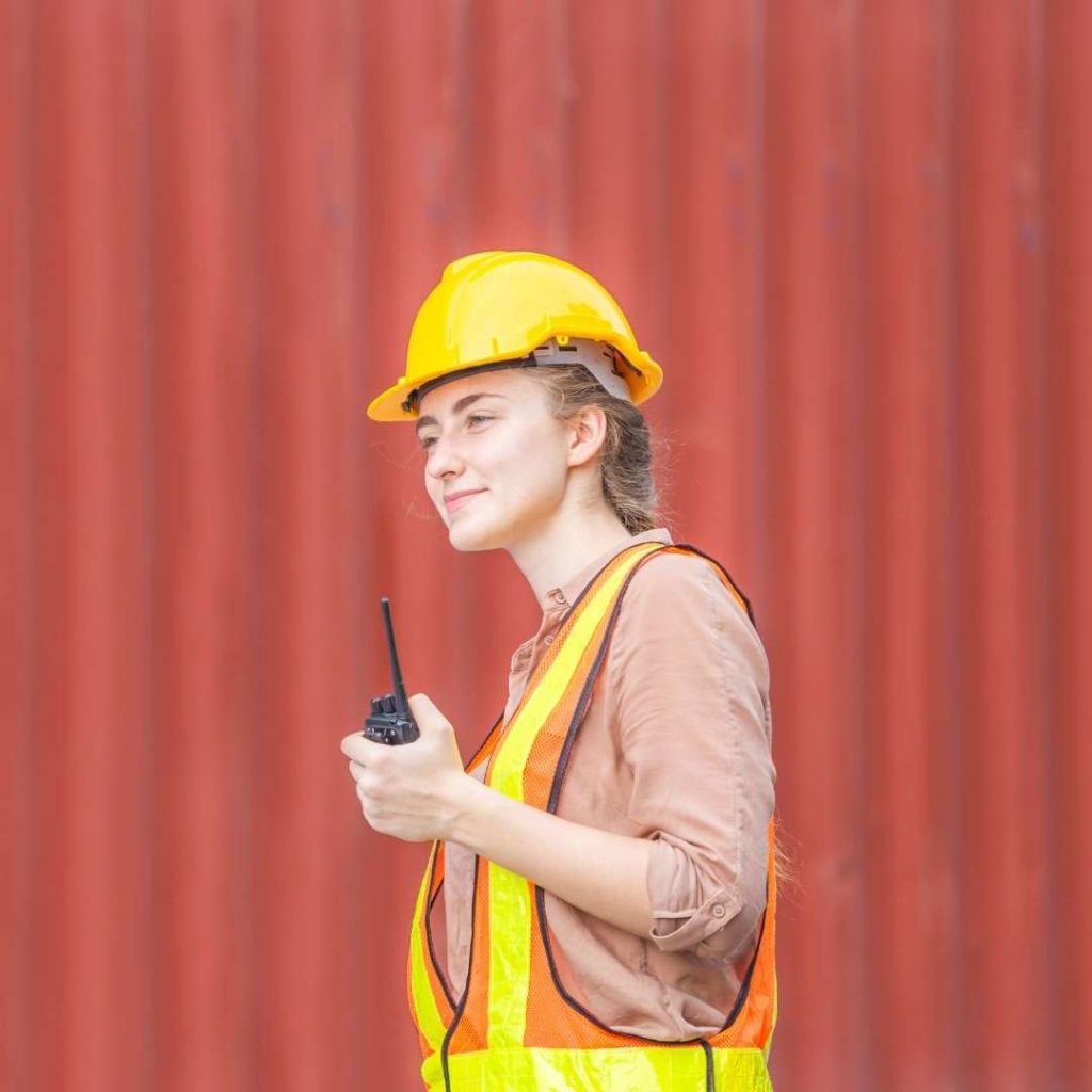 Woman in construction safety gear using walkie talkie in front of shipping container